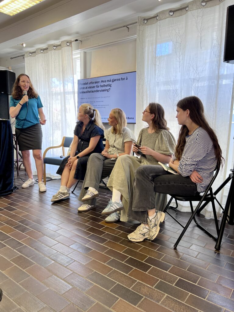 Fotografi fra en panelsamtale av fem kvinner, hvor fire sitter og en står. Kvinne som står snakker i en mikrofon.